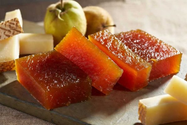 Plusieurs morceaux de pâte de coing translucides et brillantes, derrière, du queso manchego et de coings